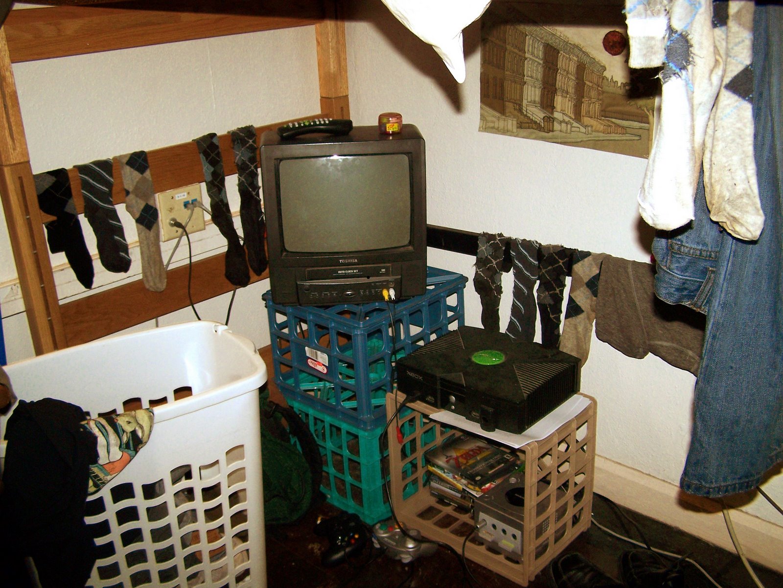 A college dorm room with a CRT on milk crates plugged into an xbox and gamecube. clothing is hung up to dry around the entertainment center.